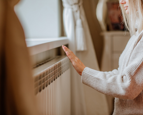 Young woman in long winter beige sweater is posing at home near the radiator