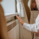 Young woman in long winter beige sweater is posing at home near the radiator